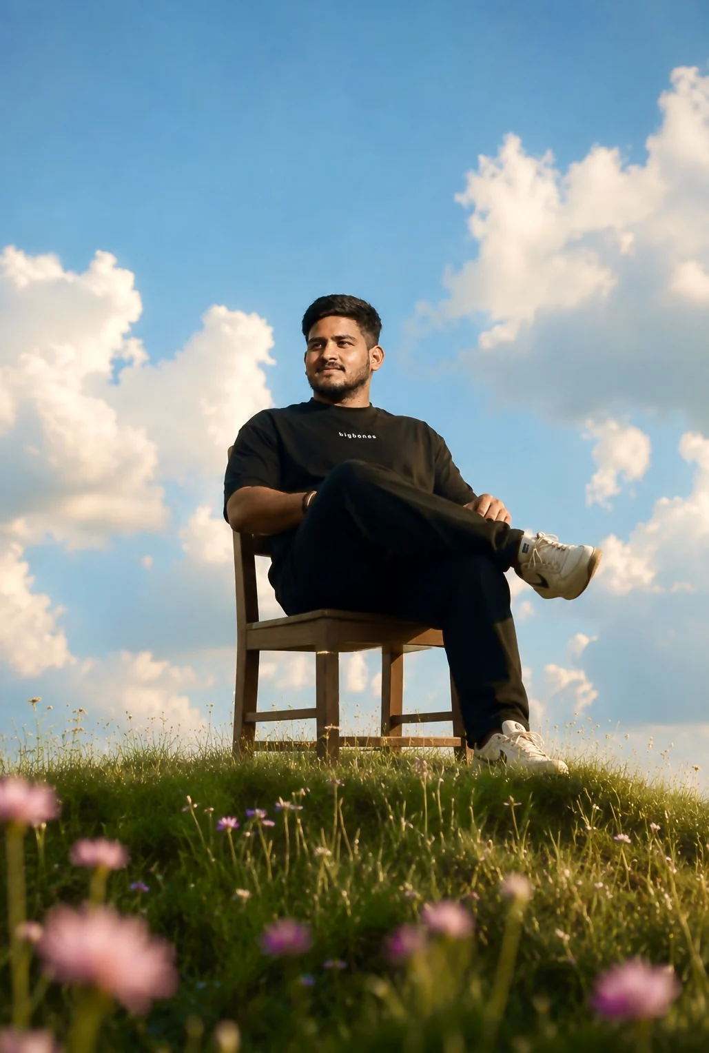 Zillionaire Honey sitting on a wooden chair on a grassy hill, dressed in black against a bright sky, wildflowers in the foreground.